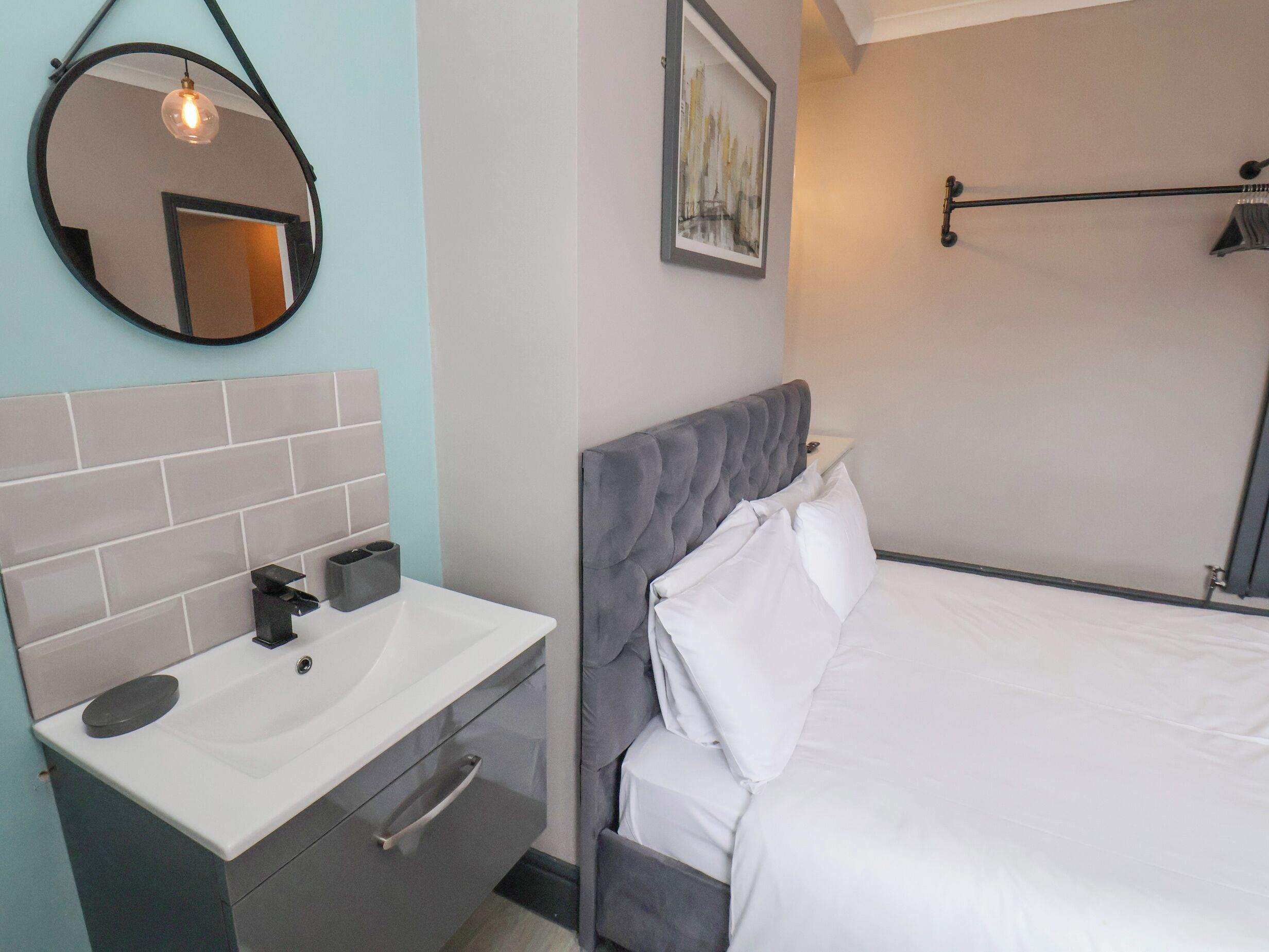 Une chambre moderne avec une tête de lit rembourrée de couleur grise, un lit blanc, un miroir rond avec une lampe suspendue, un lavabo blanc avec un meuble gris et une œuvre d’art encadrée accrochée au mur.