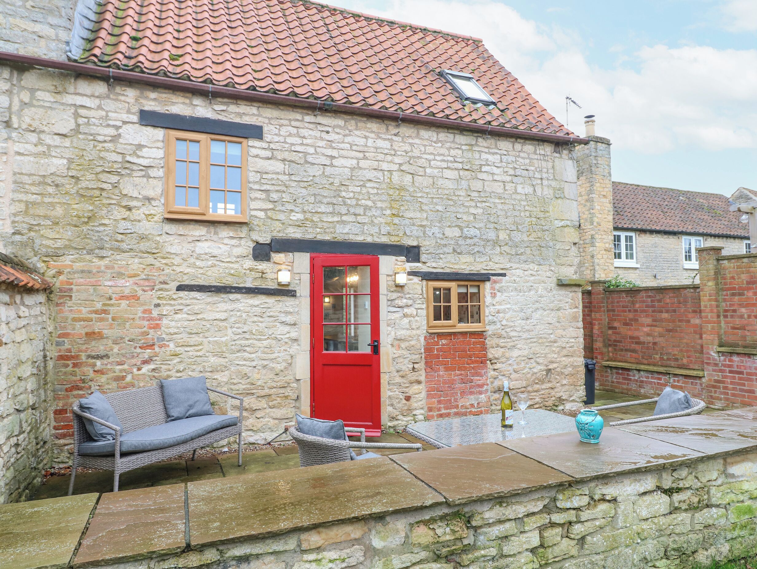 A stone building with a red door, a small window, and a brick chimney. There is a wicker sofa and a table with a wine bottle and glass.
