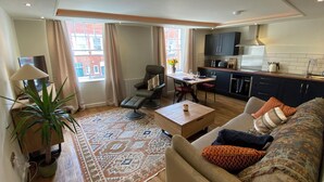 A modern living room with a sofa, armchair, and coffee table. There is a dining area with a table and chairs. The kitchen is visible with dark cabinets and a stove. - Picadore Two (Scarborough)