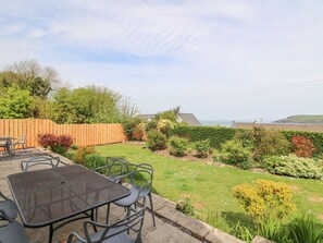 A patio with a table and chairs, a wooden fence, and a garden with various plants and a view of the sea.