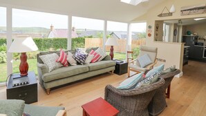 A spacious living room with a sofa, chairs, and a dining table, large windows, and a view of the countryside. - Tree Tops (Newport)