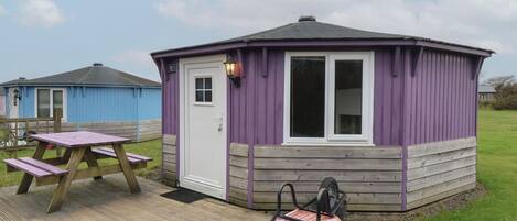 A small, single-story cabin with a purple exterior and a white door, featuring a picnic table and a wheelbarrow outside.