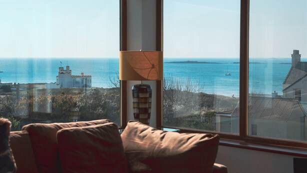 A room with a large window overlooking a coastal view, a lamp, and a sofa. - Swyn Llyr (Rhosneigr)