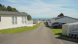 A row of mobile homes with a paved road in between, a clear sky, and a view of the ocean in the distance. - Chalet 15 at Landscove Holiday Park (Brixham)