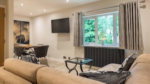 Una sala de estar moderna con un sofá, una mesa de centro de vidrio, una zona de comedor con sillas, un televisor de pantalla plana en la pared y una ventana con cortinas.