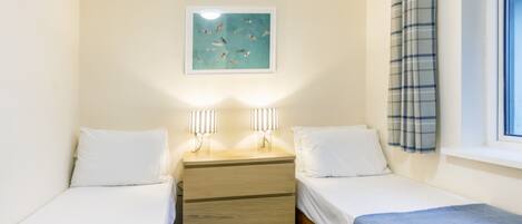 Un dormitorio con dos camas individuales, una mesita de noche con dos lámparas, un cuadro en la pared y una ventana con cortina.