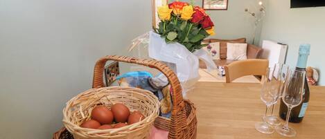 A dining area with a wicker basket containing eggs and a bouquet of flowers, two wine glasses, and a bottle of champagne on a wooden table.