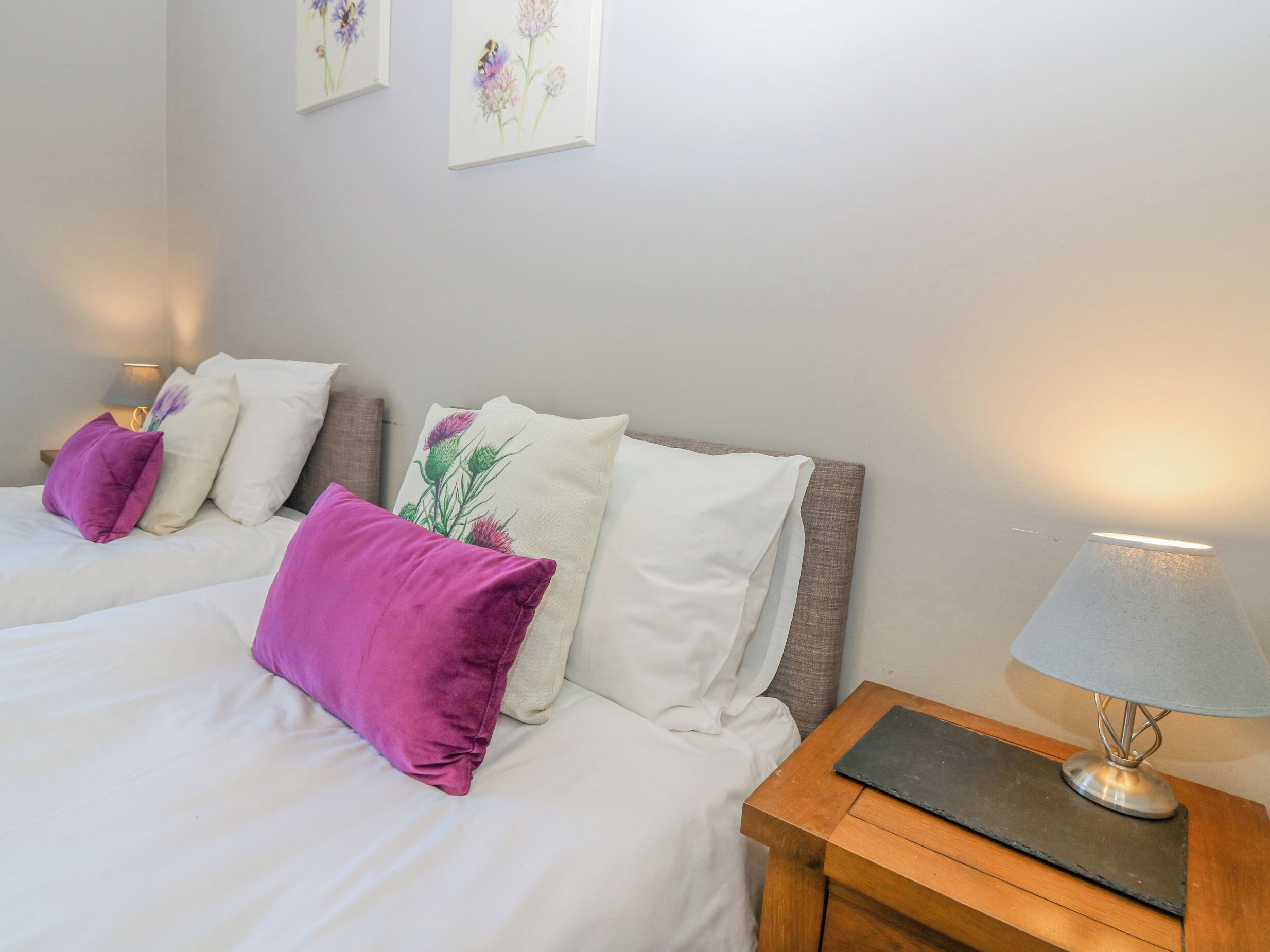 Een slaapkamer met twee bedden, elk met witte beddengoed en paars-witte kussens. Er staat een nachtkastje met een lamp en een klein zwart voorwerp op.