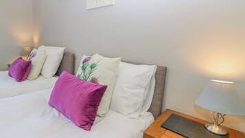 Een slaapkamer met twee bedden, elk met witte beddengoed en paars-witte kussens. Er staat een nachtkastje met een lamp en een klein zwart voorwerp op.