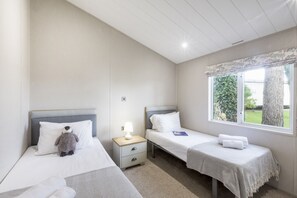 A hotel room with two single beds, a nightstand with a lamp, a window with a view of greenery, and a small table with a book. - Lodge 9 at Landscove Holiday Park (Brixham)