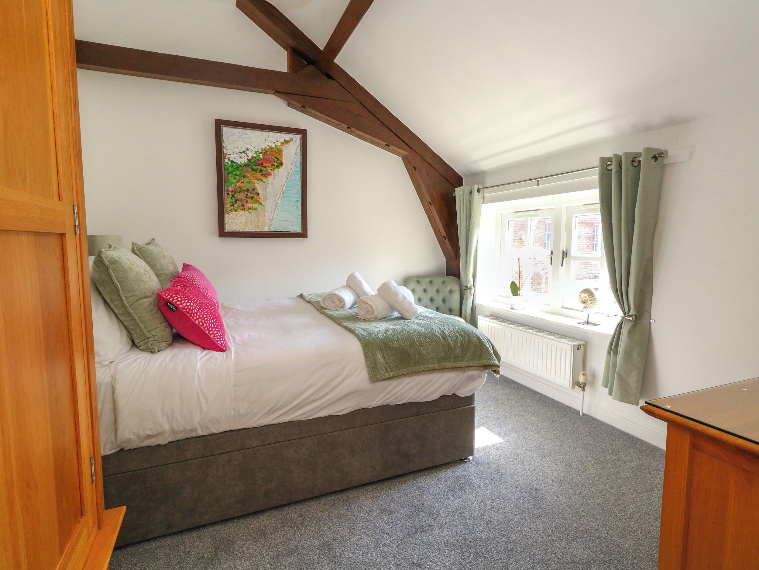A bedroom with a bed, pillows, a green blanket, a window with curtains, and a radiator.
