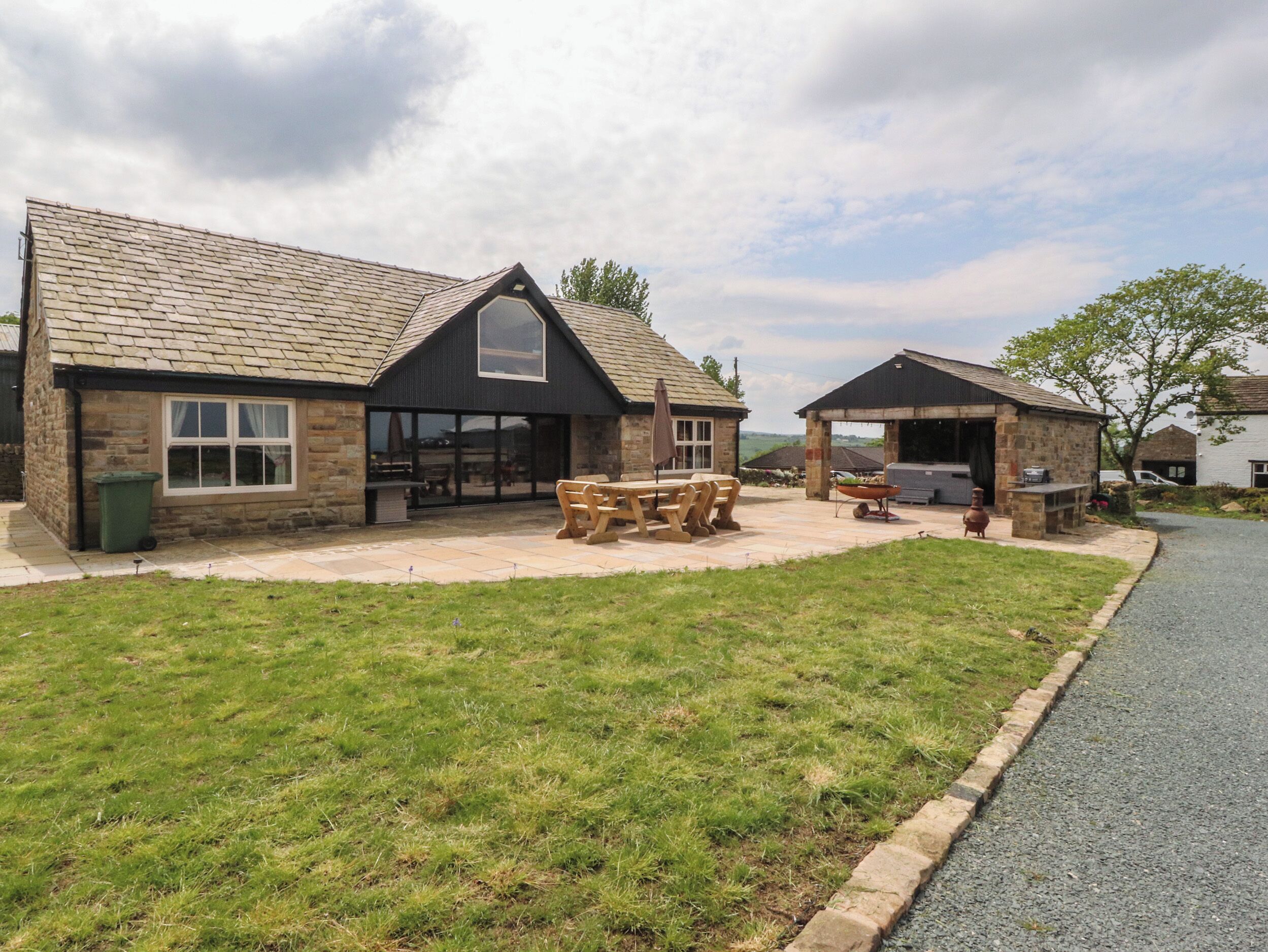 A stone building with a tiled roof, a large patio area with a wooden table and benches, and a barbecue setup.