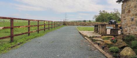 Un chemin de gravier mène à un bâtiment en pierre, bordé d’une clôture en bois d’un côté.