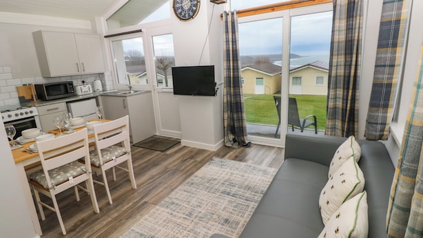 A compact kitchen with a microwave, toaster, and sink, a dining area with a table and chairs, a gray sofa with bee-patterned pillows, and a large window with plaid curtains. - Chalet 22 (Swansea)