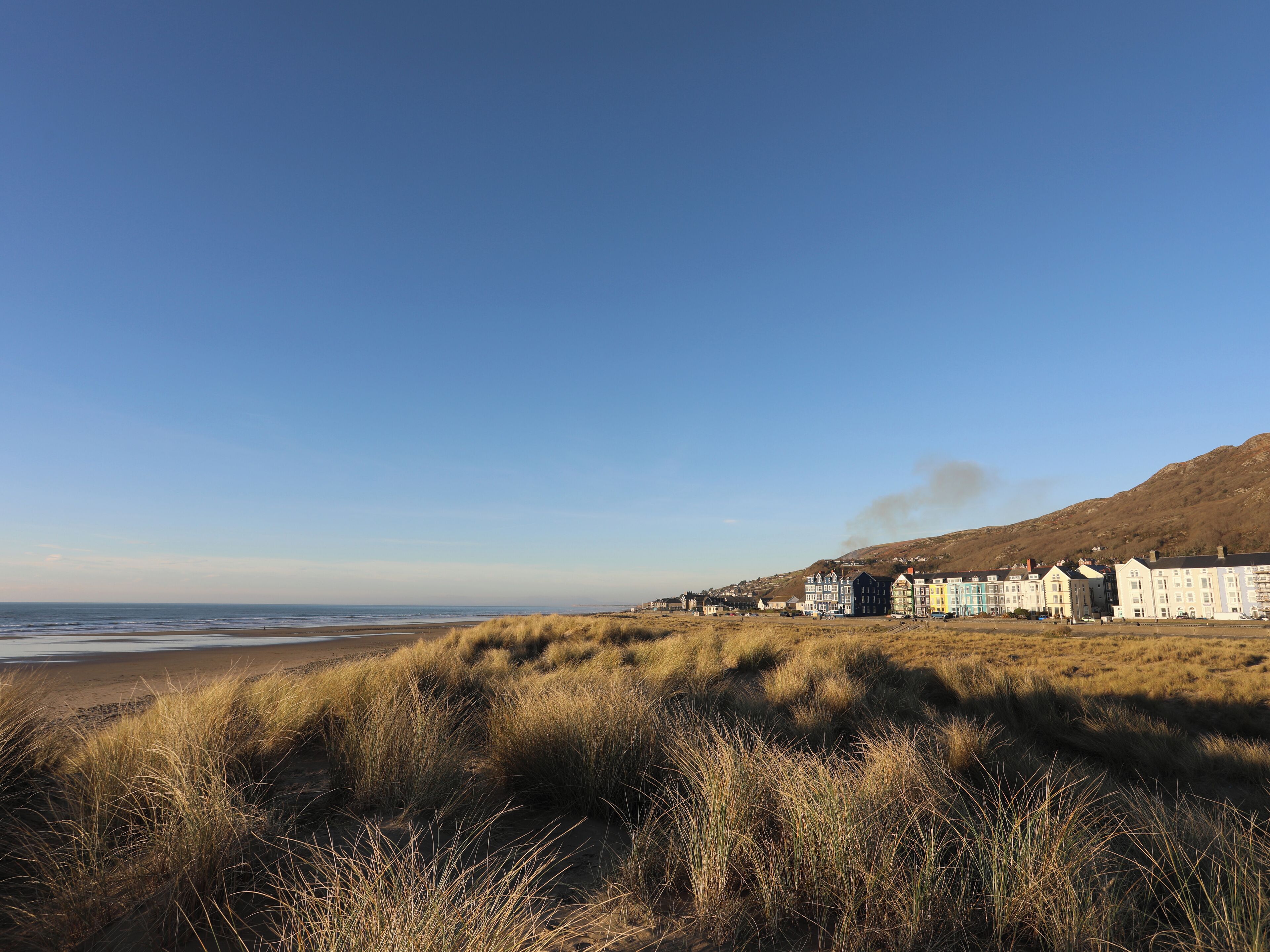A coastal town with buildings along the beach, a clear sky, and a hill in the background.