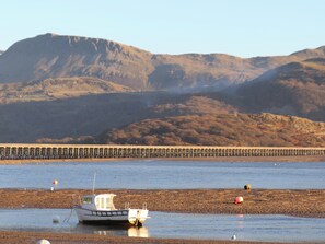 A boat on a pebbly shore with a pier extending into the water and hills in the background. - Sandcastles (Barmouth)