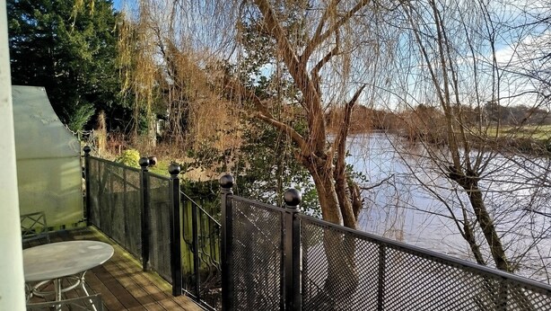 A deck with a table and chairs overlooking a river, surrounded by trees and a fence. - River Cottage (Derby)