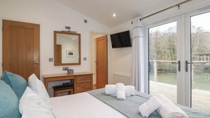 Une chambre à coucher comprenant un lit, une commode, un miroir, un téléviseur et un balcon avec vue sur des arbres et une étendue d’eau.