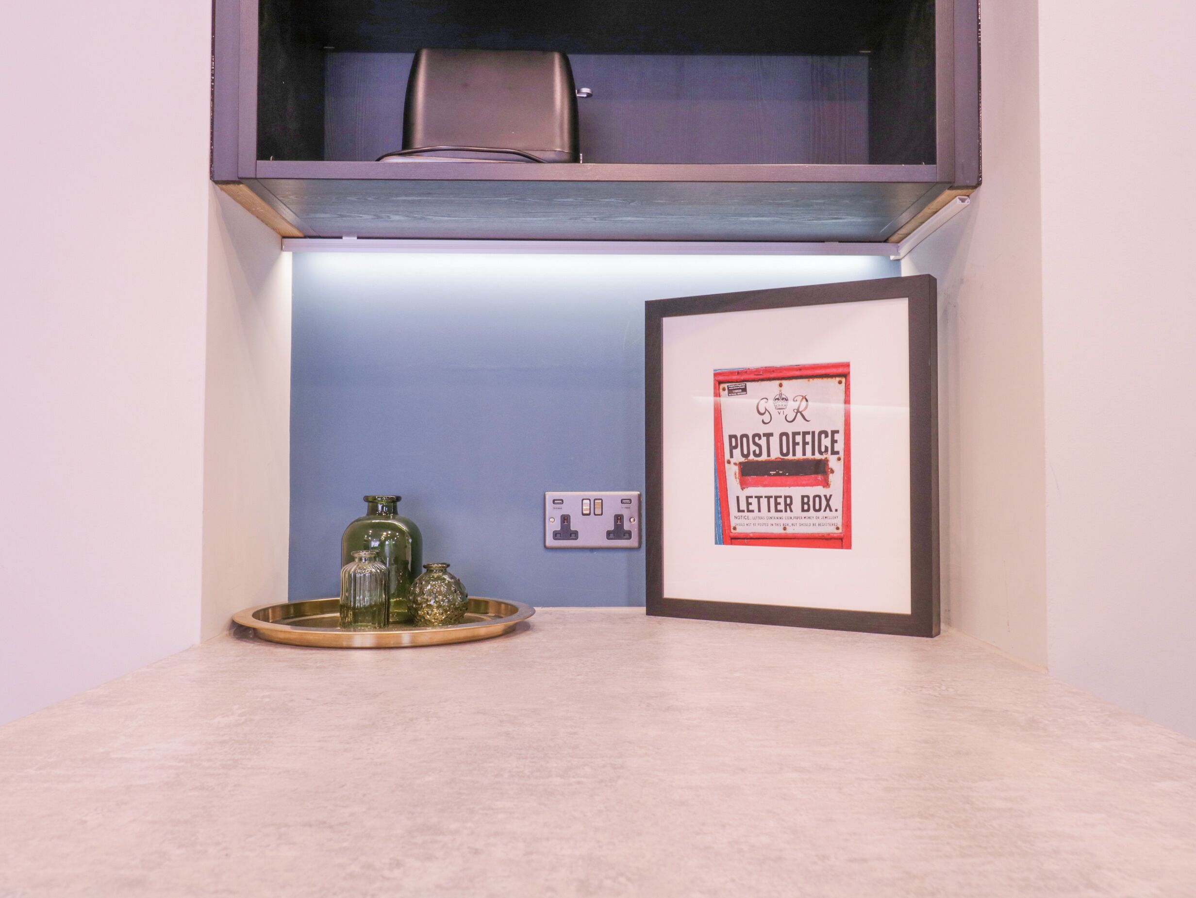 A framed sign for a post office letter box, a small table with decorative glass bottles, and a power outlet on a wall.