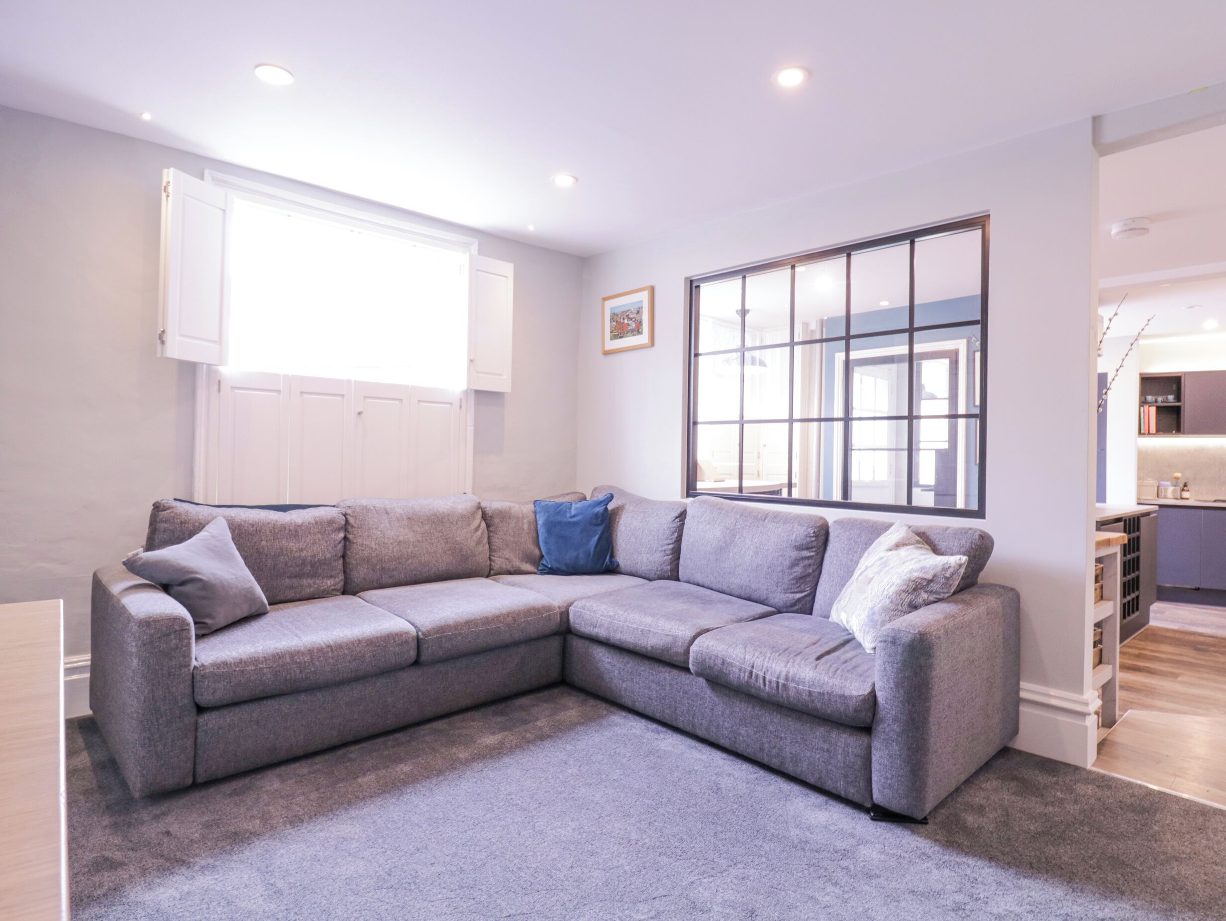 A modern living room with a grey sectional sofa, a large window, and a kitchen area visible through an open door.