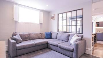 A modern living room with a grey sectional sofa, a large window, and a kitchen area visible through an open door.