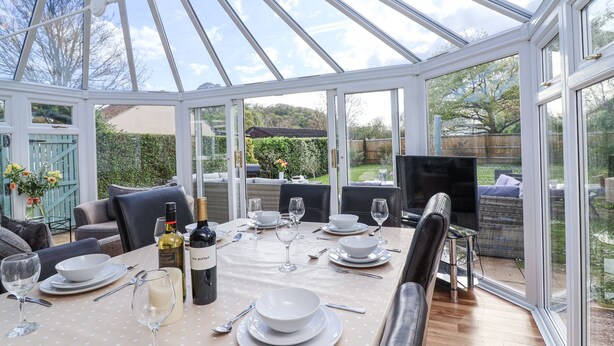 A conservatory with a glass roof, a dining table set for six, and a cozy seating area with cushions.