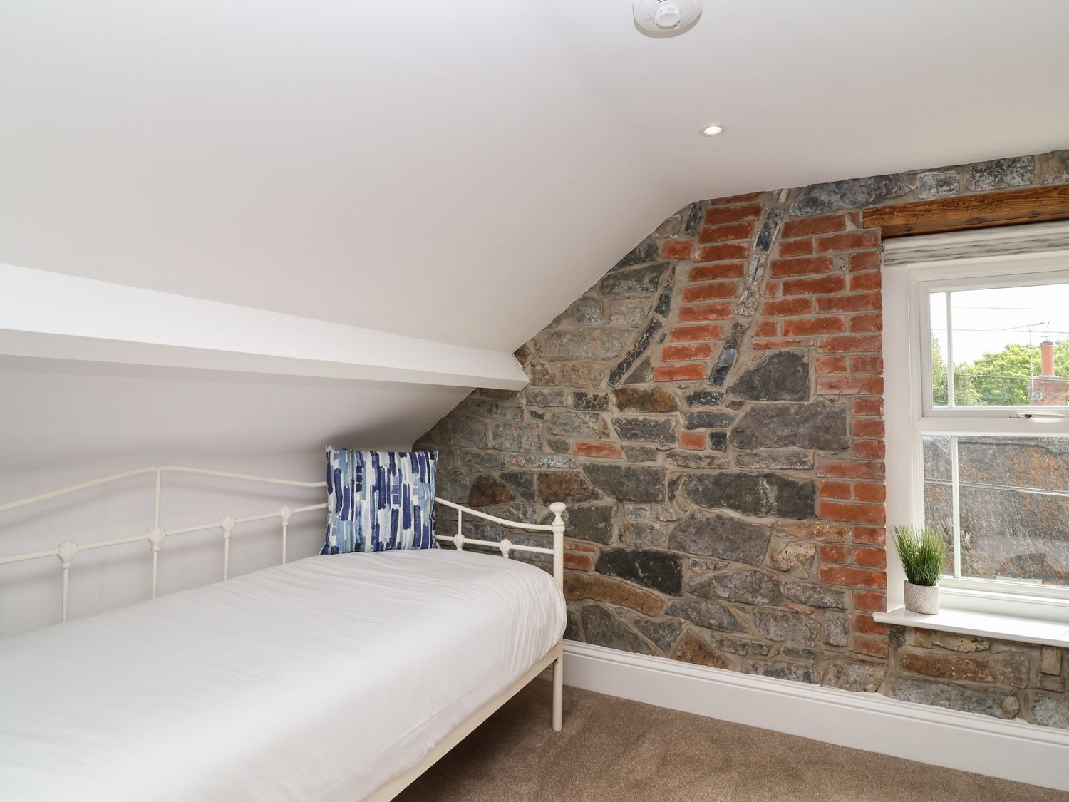 A bedroom with a sloped ceiling, a white bed, a stone wall, and a window with a plant.