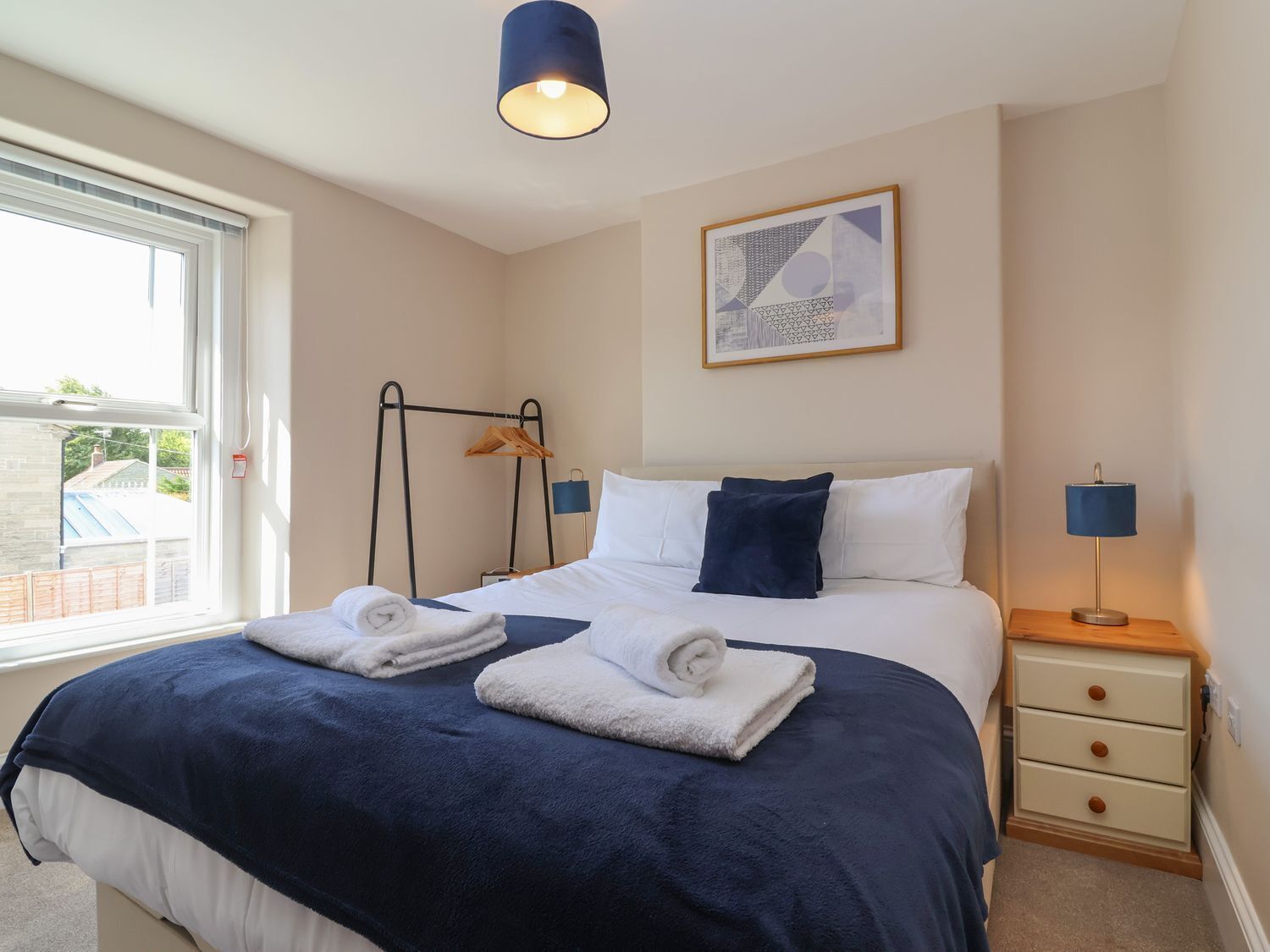A neatly made bed with white and navy bedding, a bedside lamp, a framed abstract artwork on the wall, and a window with a view of the outside.