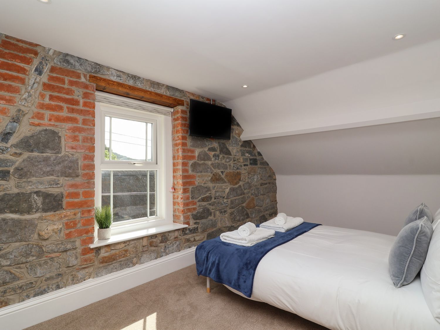 Un dormitorio con pared de piedra, una ventana, una cama con manta azul y un televisor de pantalla plana montado en la pared.