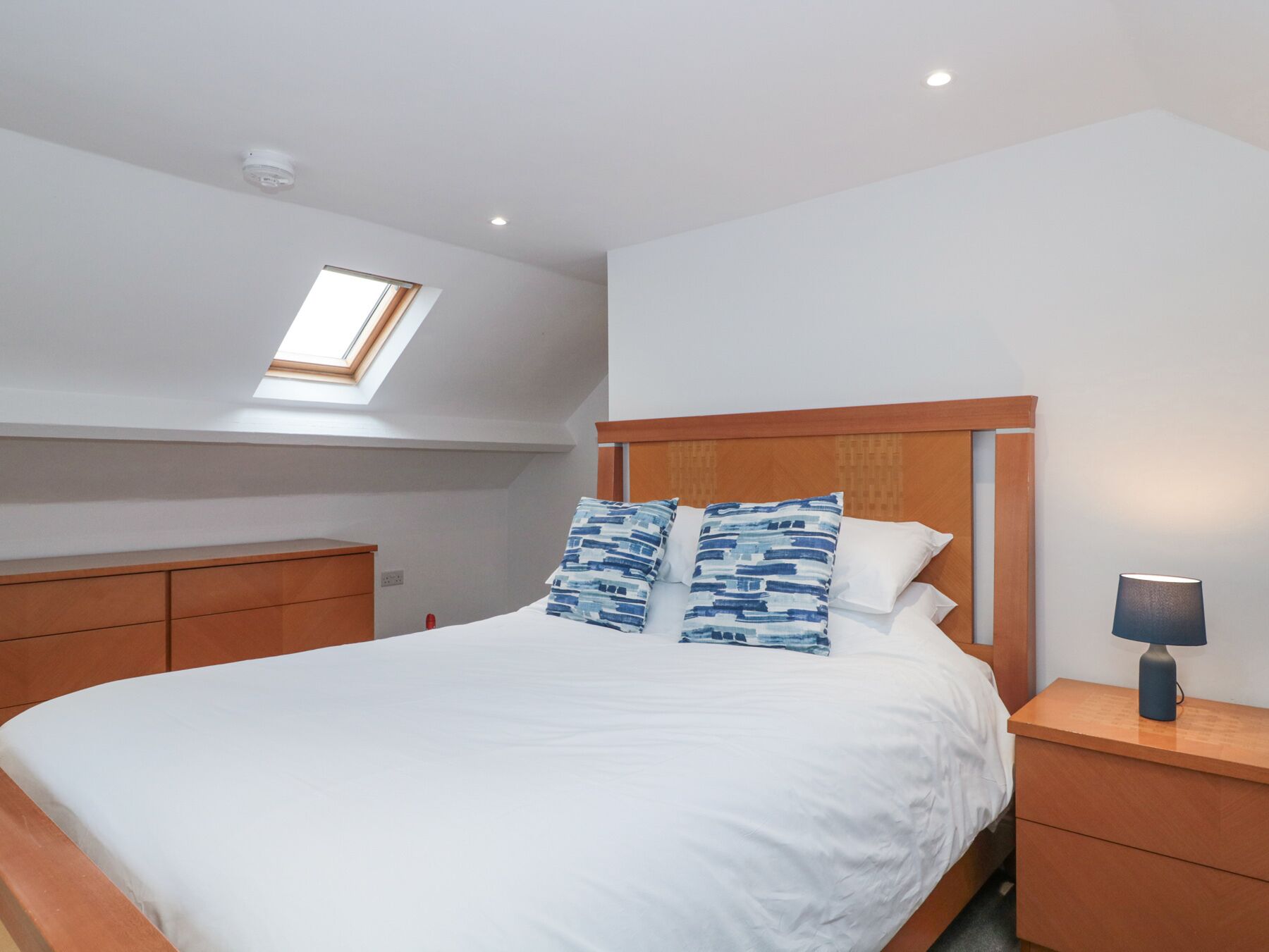Un dormitorio con tragaluz, cama con ropa de cama blanca y almohadas azules, cabecera de madera y mesita de noche con lámpara.