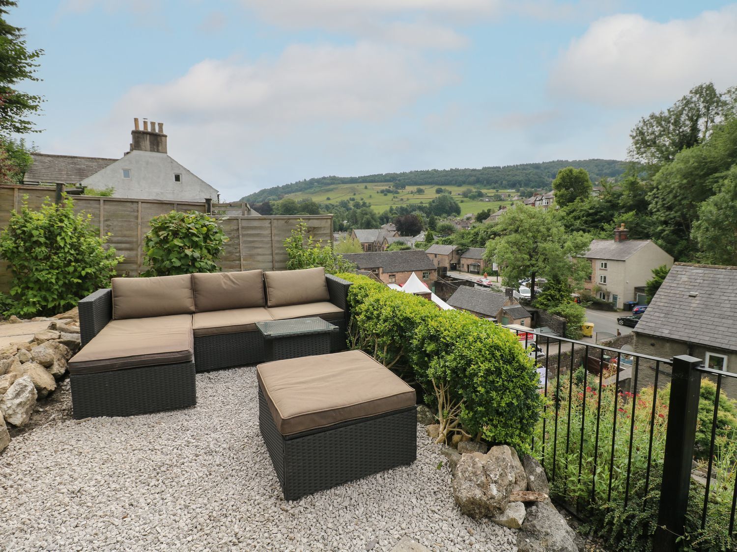 An outdoor seating area with a sofa and ottoman, surrounded by gravel and greenery, overlooking a village with houses and hills in the background.