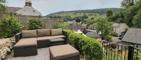 An outdoor seating area with a sofa and ottoman, surrounded by gravel and greenery, overlooking a village with houses and hills in the background.