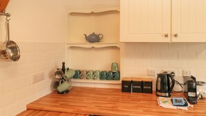 A kitchen with white cabinets, a wooden countertop, a kettle, a coffee maker, and a shelf with mugs and a teapot.