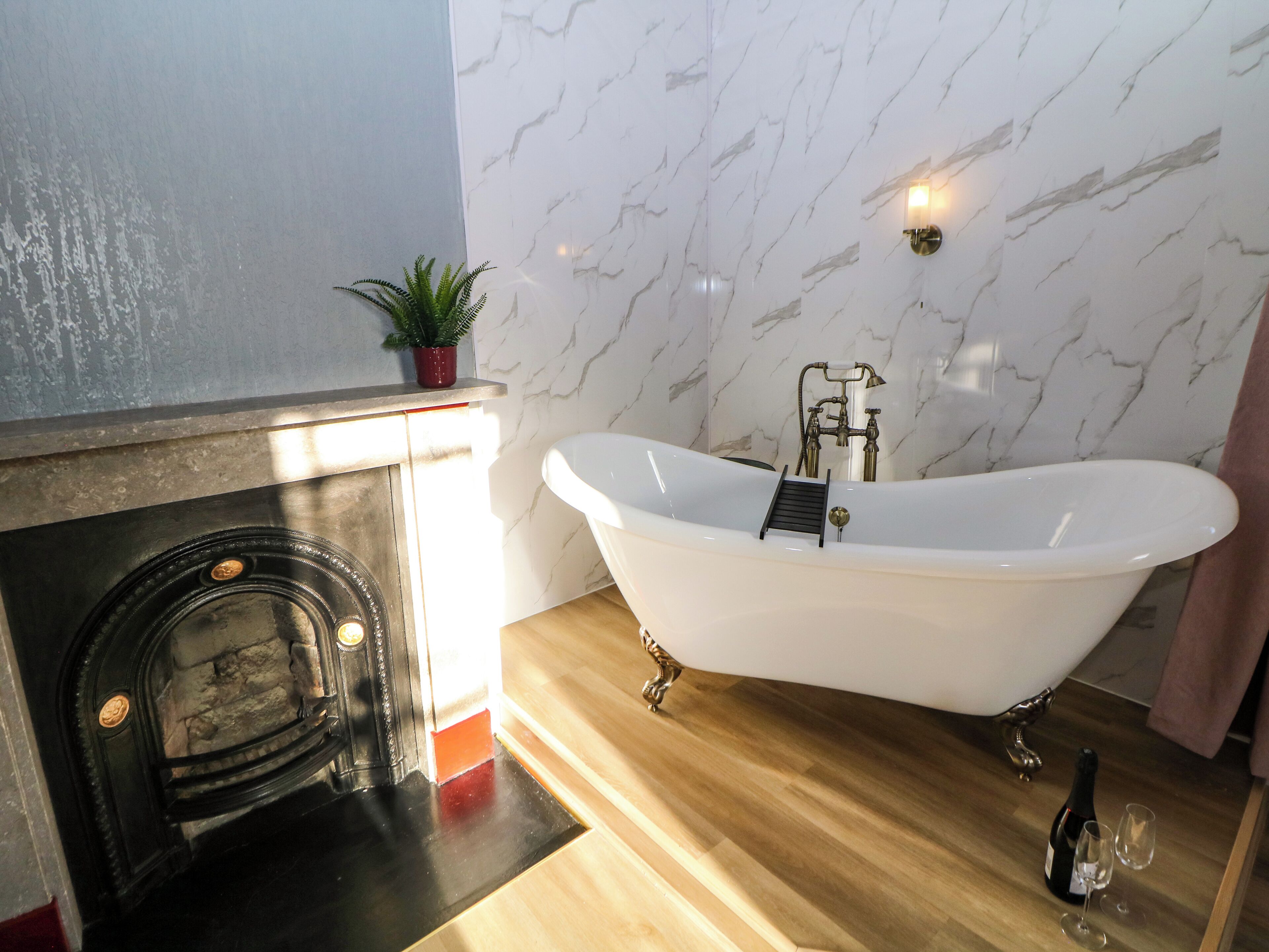 A bathroom with a freestanding bathtub, a marble wall, a small potted plant, and a bottle of champagne on the floor.