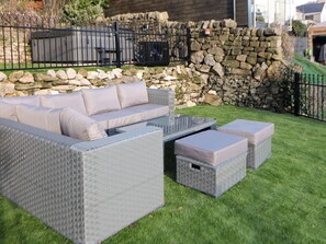 Un espacio de descanso al aire libre con asientos de mimbre gris, cojines, una mesa de centro de vidrio y cajas de almacenamiento.