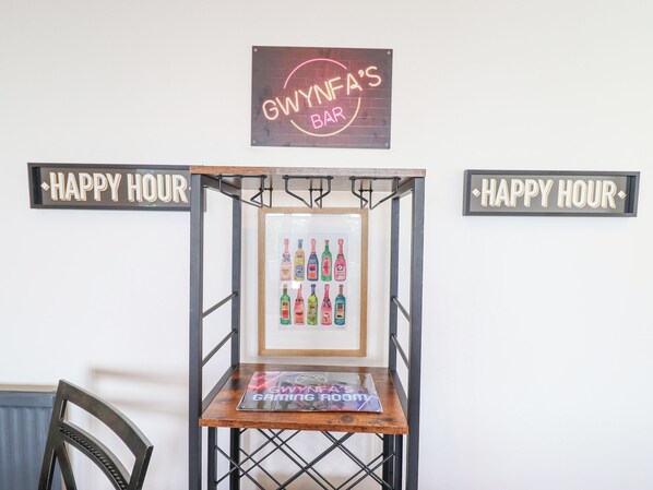 A bar area with a neon sign reading "GWYNA'S BAR" and two signs saying "HAPPY HOUR". A framed artwork of colorful bottles is displayed on a wooden table. - Gwynfa (Barmouth)