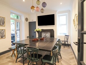 Une salle à manger moderne avec une grande table rectangulaire, plusieurs chaises, un téléviseur à écran plat fixé au mur et une cheminée avec un manteau en marbre.