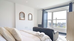 A modern bedroom with a large bed, a dark sofa, and a view of the city through a large window. - Gwynfa (Barmouth)