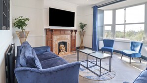 A modern living room with a fireplace, blue sofas, a glass coffee table, and a large window with blue curtains. - Gwynfa (Barmouth)