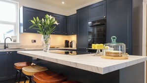 A modern kitchen with dark cabinets, a marble countertop, a vase with flowers, and a cake on a stand. - Dolfor (Cemaes Bay)