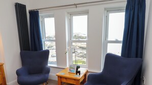 A room with two blue armchairs, a small wooden table, and a view of a coastal area through large windows. - Dolfor (Cemaes Bay)