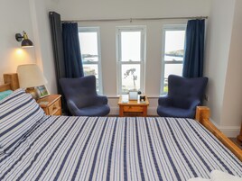 Habitación de hotel con una cama grande, dos sillones, una mesita de noche con lámpara y vistas al mar a través de dos ventanas.