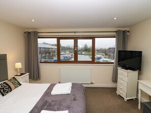 A hotel room with a bed, a TV, a dresser, a window with a view of boats and buildings, and a radiator. - 4 Teal House (Carnforth)