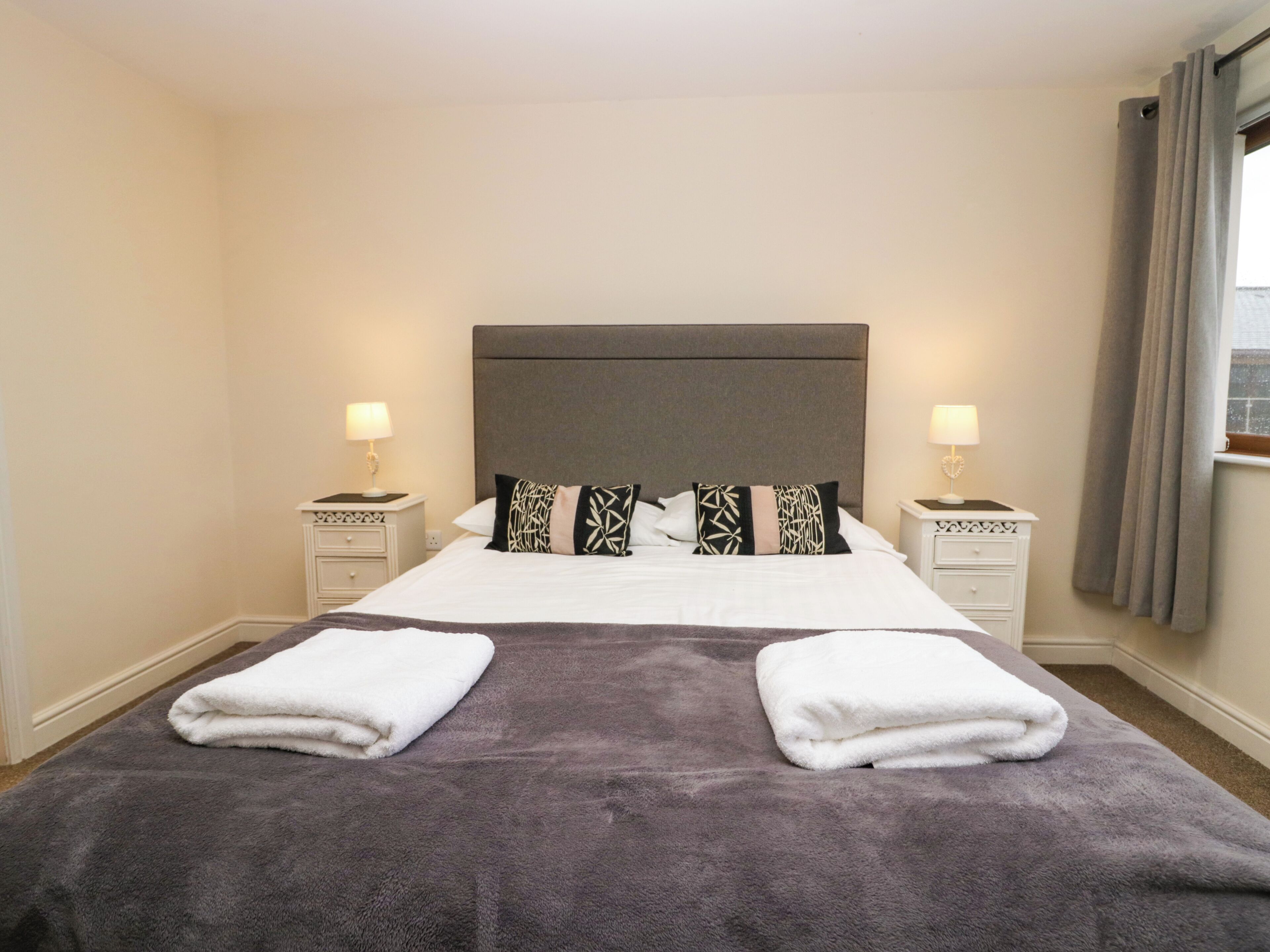 A hotel room with a large bed, two nightstands, a grey headboard, and a window with curtains.
