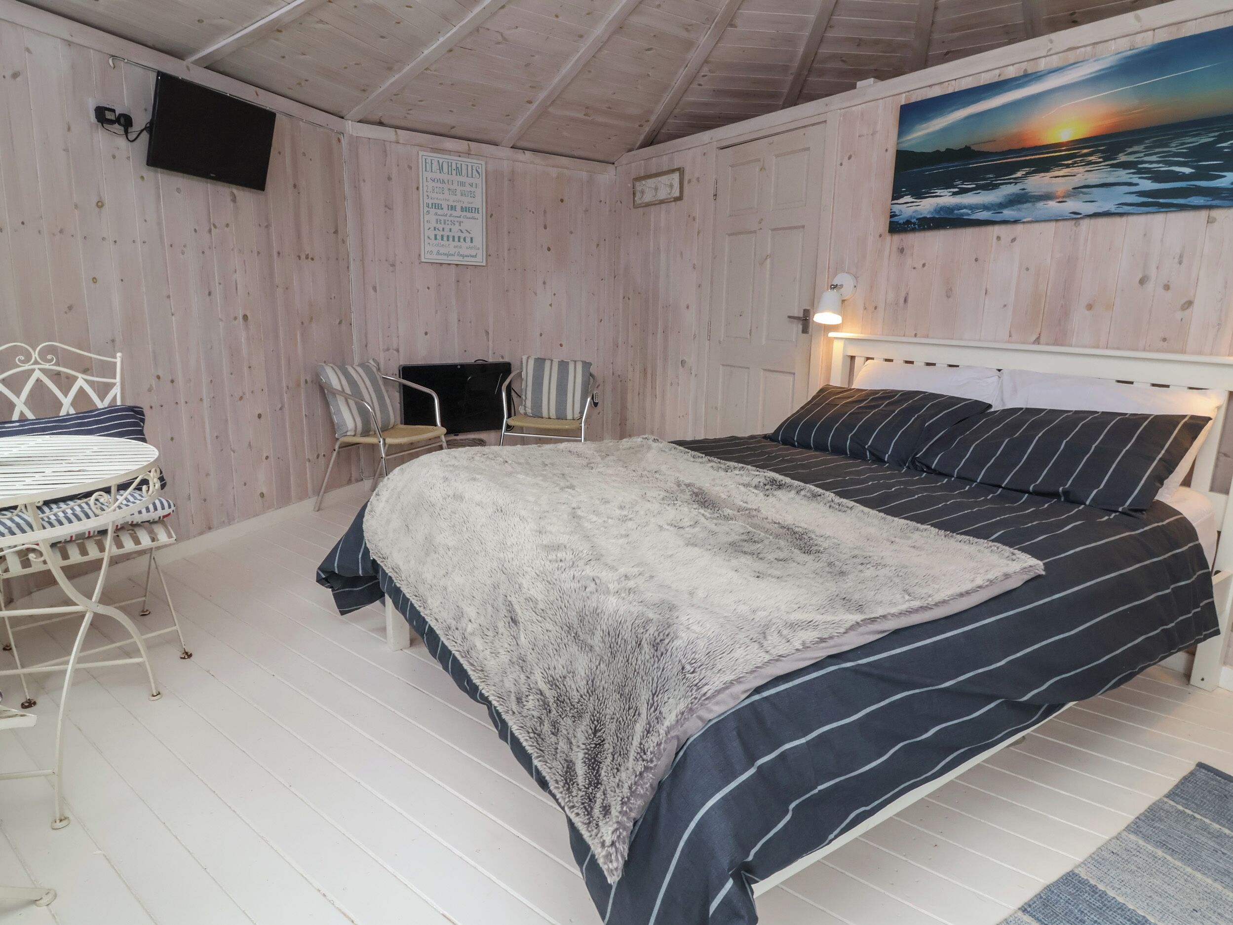 Um quarto aconchegante com cama, poltrona, mesinha e uma pintura de pôr do sol.