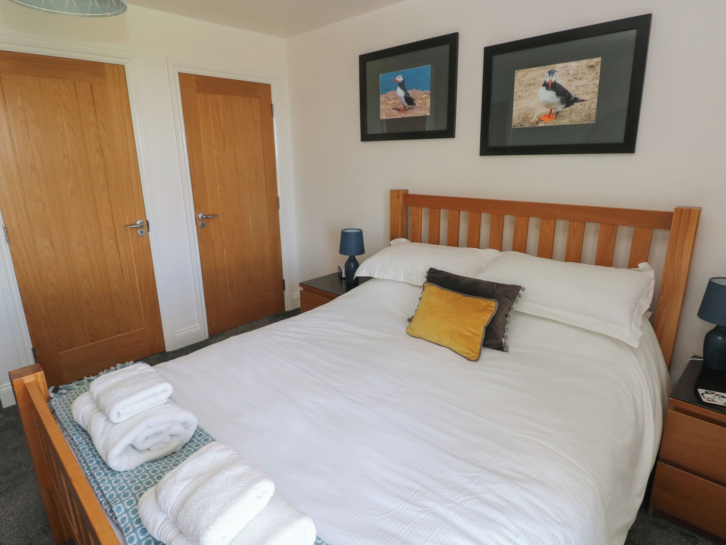 A neatly made bed with white linens and a single yellow pillow, a wooden headboard, and two framed pictures of puffins on the wall.