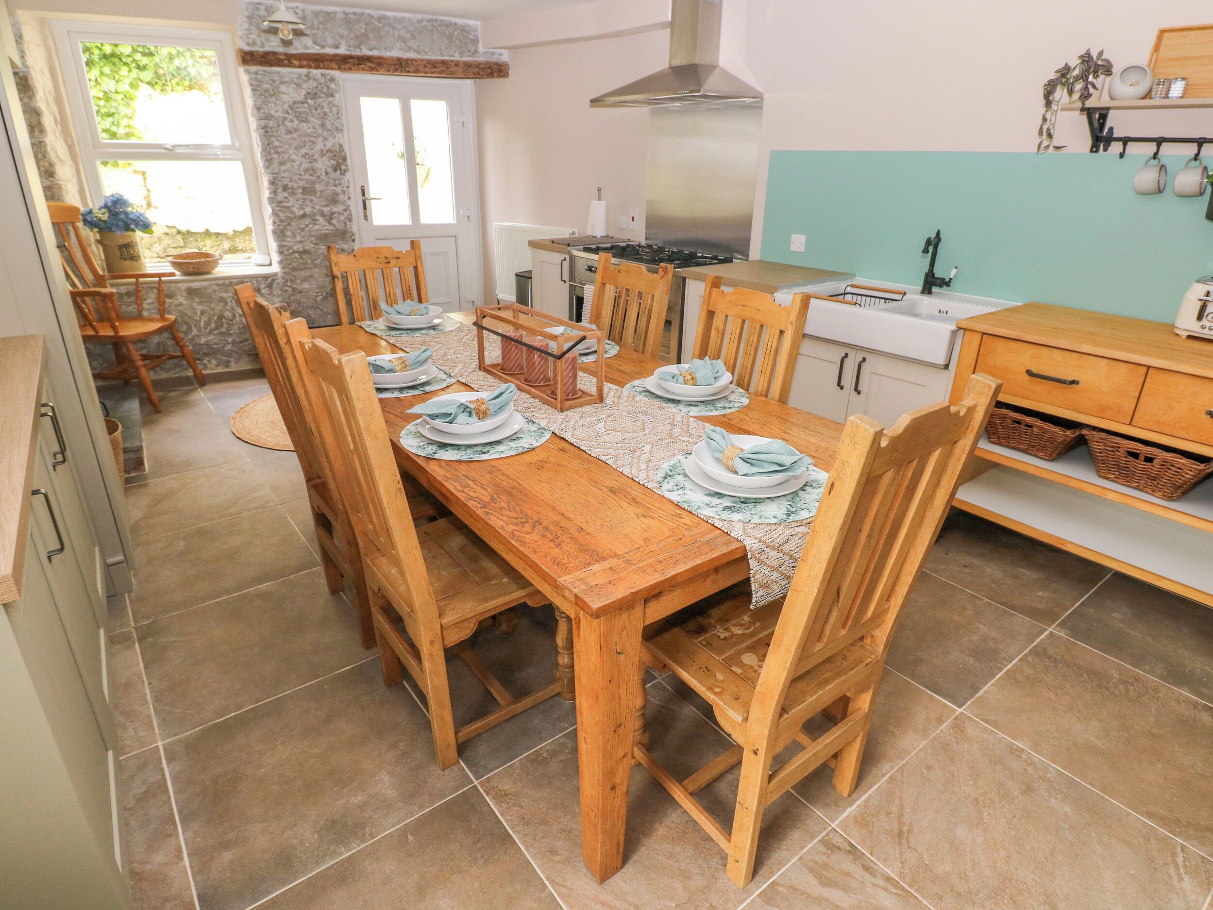 Un comedor con una mesa de madera dispuesta para seis personas, rodeada de sillas. Se ve una cocina con estufa y lavaplatos al fondo.