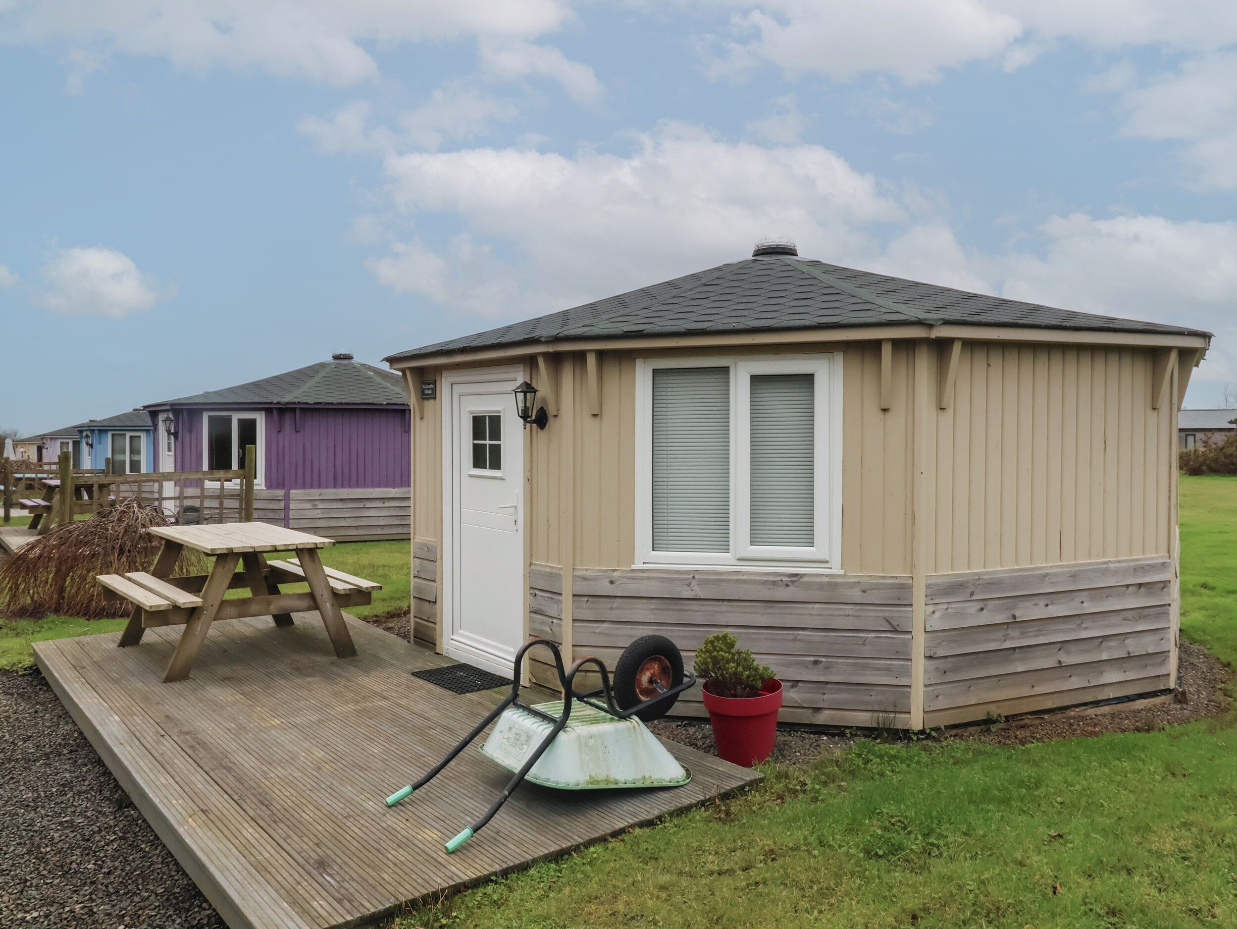 Une petite cabane en bois avec une porte, des fenêtres et un espace patio comprenant une table et une brouette.