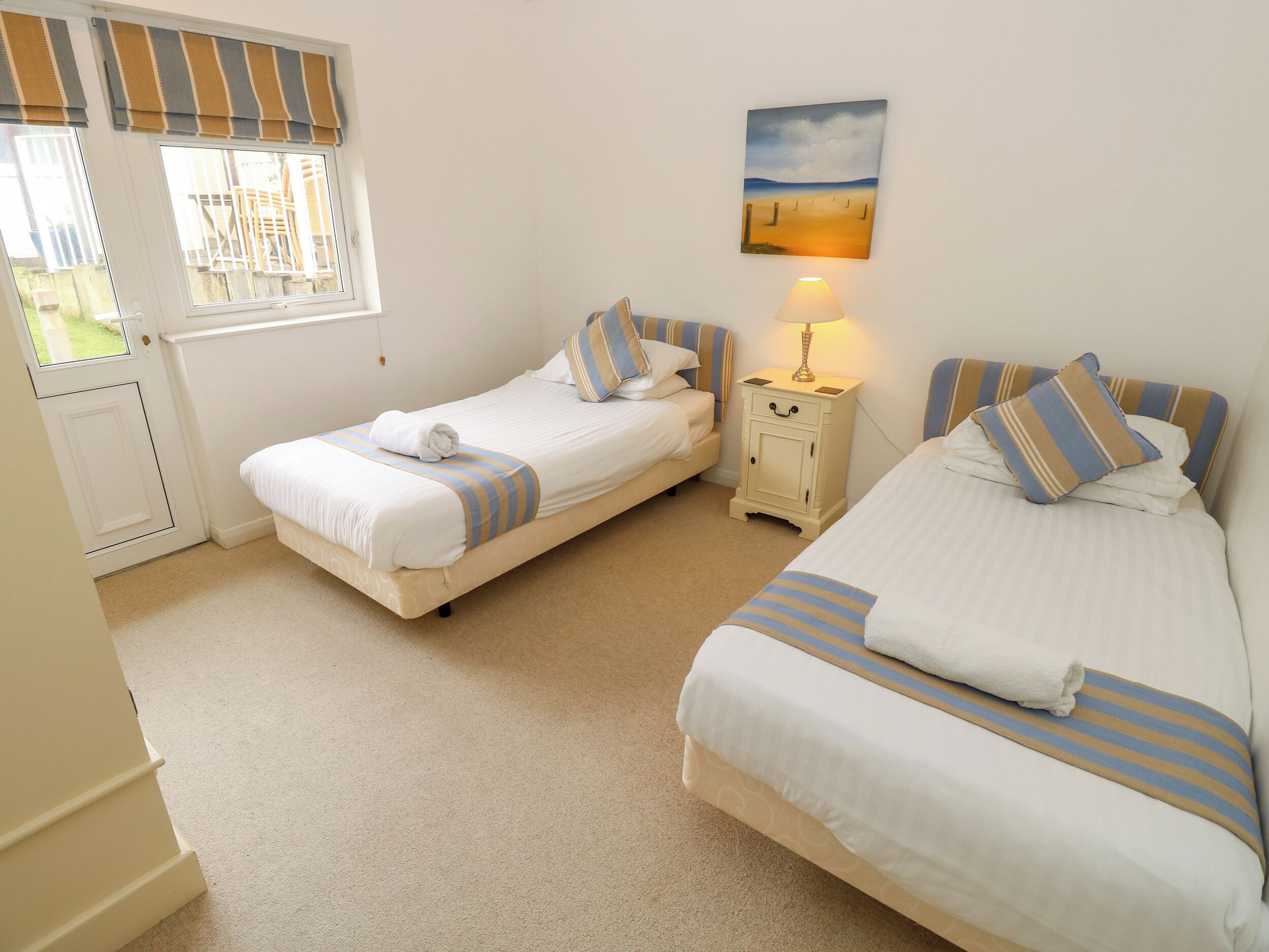 A bedroom with two beds, a nightstand with a lamp, a painting on the wall, and a door with a view of the outside.