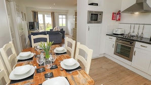 A dining area with a wooden table set for four, white chairs, and a kitchen with white cabinets, a microwave, and a stove. - Munday H2 (Yarmouth)